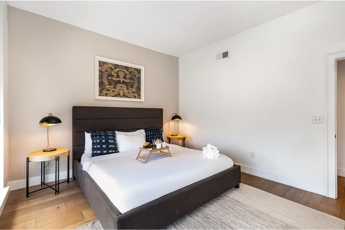 A cozy bedroom with a neatly made bed, flanked by side tables and lamps. Inviting artwork and soft lighting create a restful and intimate atmosphere, perfect for unwinding after a busy day.