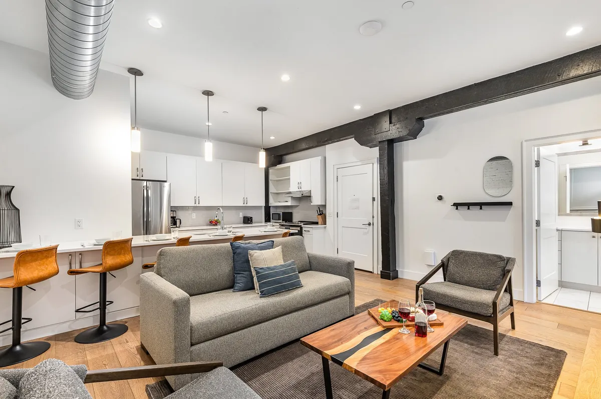 The image showcases a stylish, contemporary living area featuring a plush sofa adorned with cozy cushions, a chic wooden coffee table perfect for gathering, and a sleek kitchen with bar stools that invite conversation—all bathed in warm, inviting light.