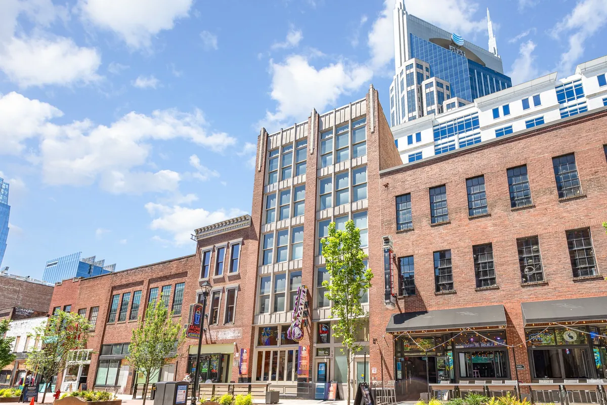 A vibrant exterior street scene featuring a historic brick building amidst modern skyscrapers, leafy trees, and an inviting outdoor seating area. The mood is lively and social, suggesting easy access to local dining, shopping, and people-watching.