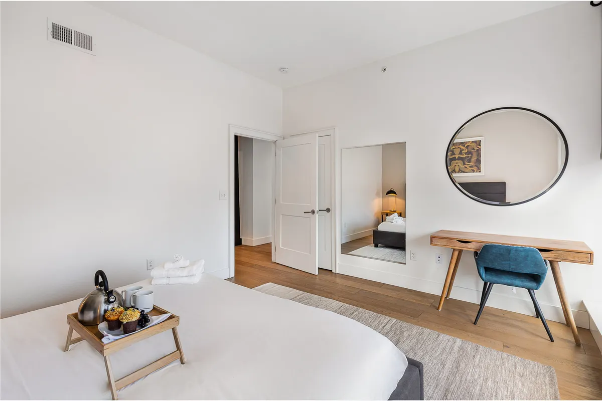 A serene bedroom scene with a king-size bed dressed in crisp white linens and a decorative tray with tea and snacks. A stylish round mirror hangs above a wooden desk and blue chair, emphasizing the room's dual purpose as a restful retreat and practical workspace.