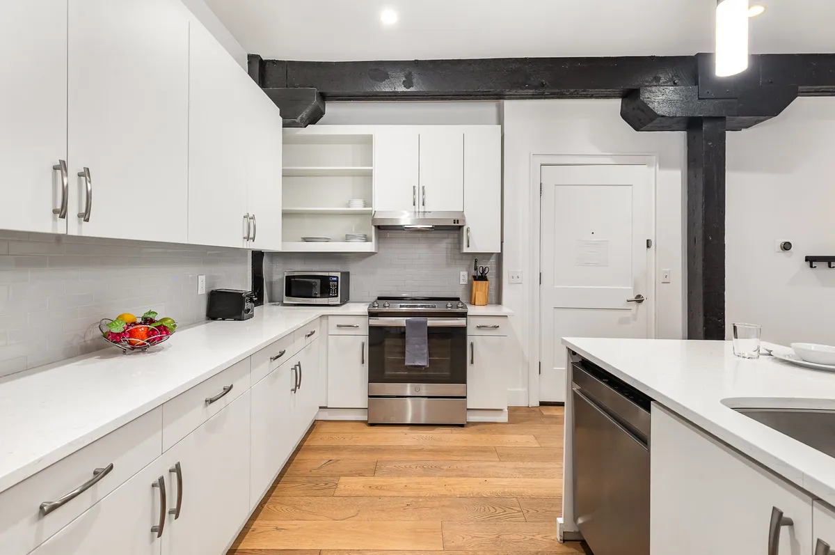 The image features a functional kitchen with a prominent island, white cabinetry, stainless steel appliances, and warm wooden flooring. The arrangement suggests space for meal prep, dining, and socializing, supporting an atmosphere of togetherness and ease.