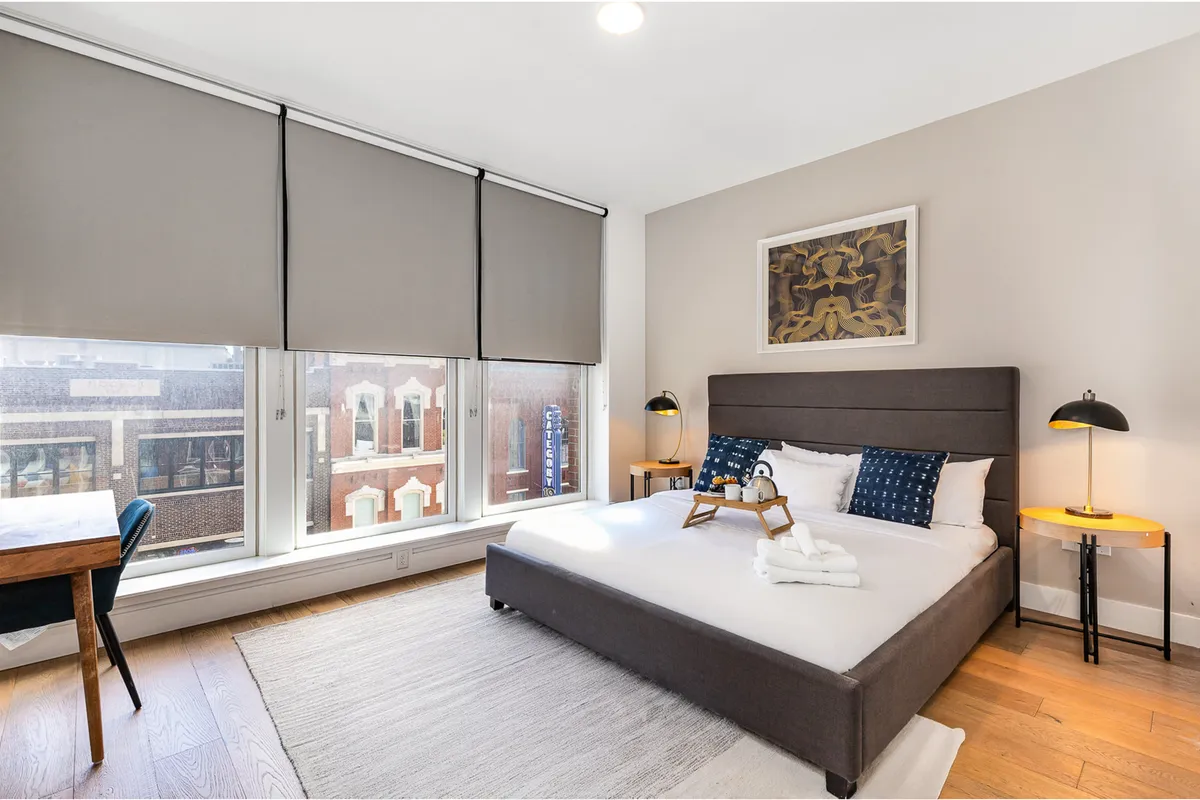 A warm, sunlit bedroom with a king bed as the focal point, accented by plush bedding, large windows with blackout shades, and eye-catching artwork. A small table with a chair sits nearby, providing a comfortable workspace with city views.