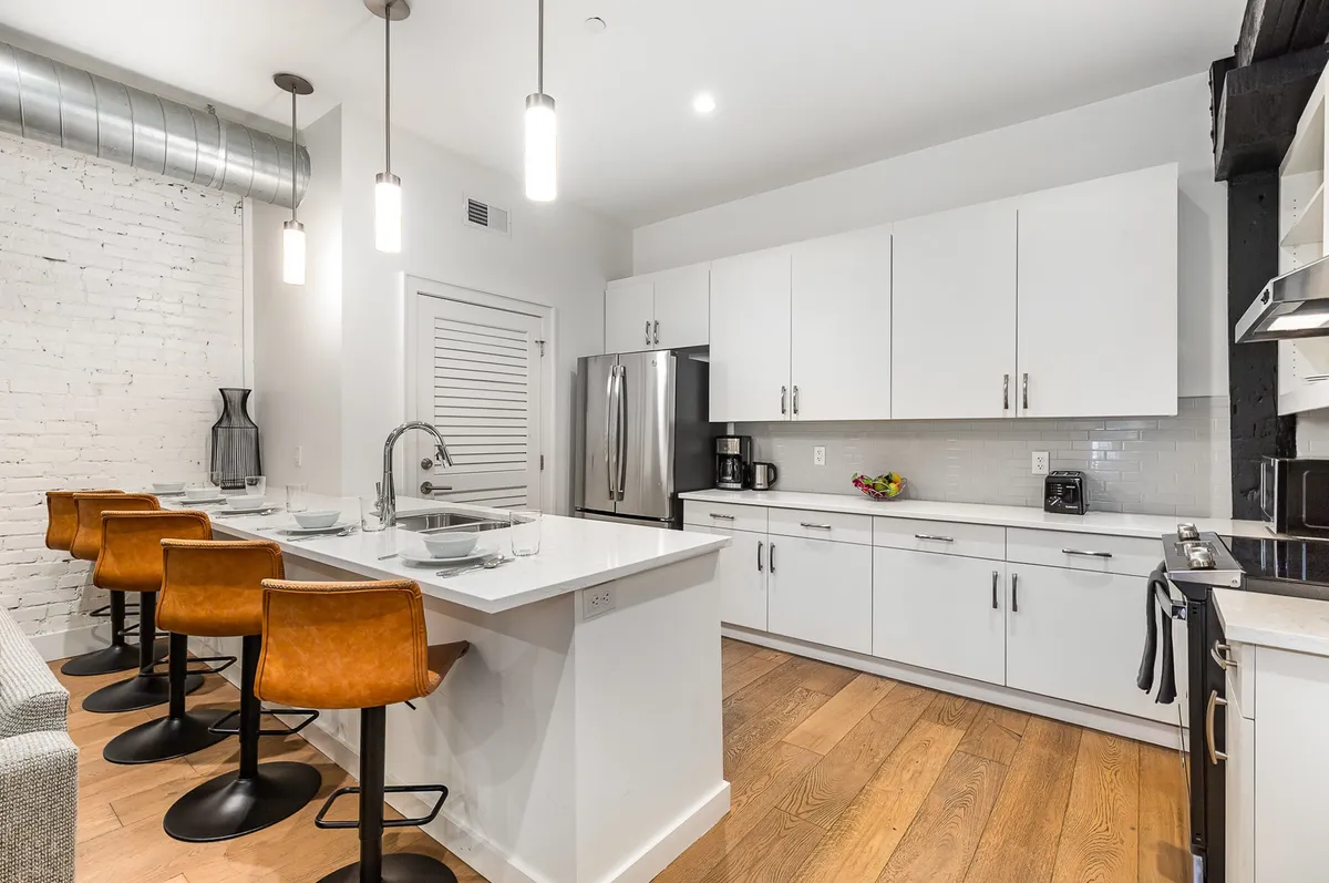 The photo centers on a spacious, contemporary kitchen with a prominent island that seats several people on bar stools. Stainless steel appliances shine against white cabinetry, and an exposed brick wall adds urban character. The space is clearly designed for both meal preparation and group gatherings, supporting activities from cooking to socializing.