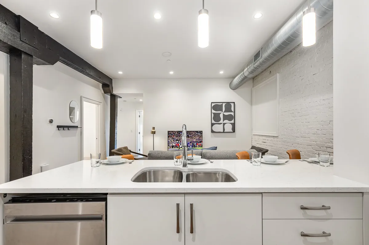 An inviting open-concept living area with a kitchen island in the background, stainless steel appliances, a comfortable sofa, and vibrant artwork on the wall. The space is sunlit and seamlessly blends the living and kitchen zones, highlighting the airy, communal atmosphere.